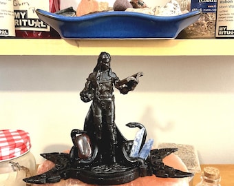 May include: A black figurine of a man holding a book, standing on a black base with a red stone and blue crystals. The figurine is on a pink salt rock.