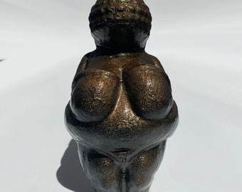 May include: A bronze-coloured figurine of a woman with exaggerated features, including large breasts and a rounded belly. The figurine is made of a textured material and has a smooth, polished finish.