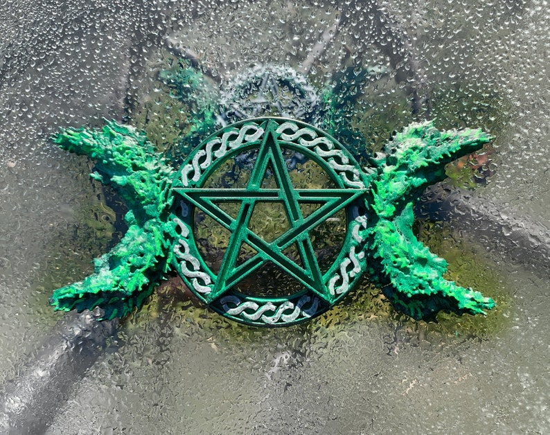 May include: Green pentagram with a triple moon symbol on either side, surrounded by a Celtic knot design. The symbol is made of a green material and is set against a blurry background.