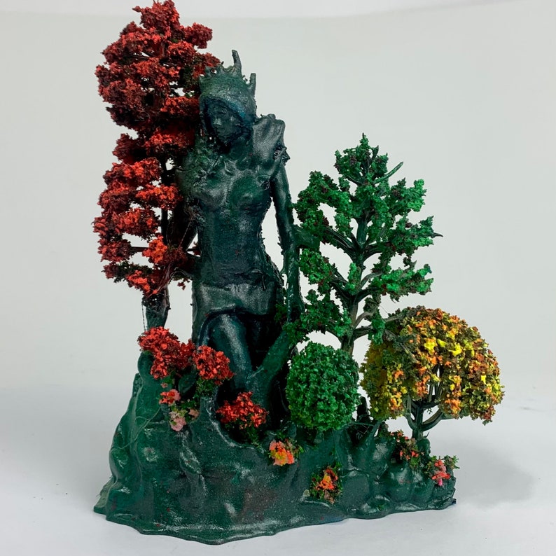May include: A 3D printed miniature scene featuring a female figure standing in a forest setting. The figure is painted in a dark green color and has a crown on her head. The trees are painted in shades of red, green, and yellow.