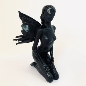 May include: A black fairy figurine with wings, kneeling with its hands clasped in front of it. The fairy has a crescent moon on its forehead.