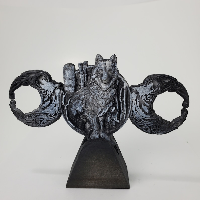 May include: A black and silver 3D printed sculpture of a wolf standing on a crescent moon. The wolf is surrounded by a decorative design of leaves and vines. The sculpture is mounted on a black wooden base.
