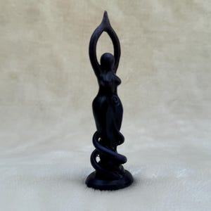 May include: Black figurine of a woman standing with her arms raised above her head, with a snake wrapped around her legs. The figurine is on a black base.
