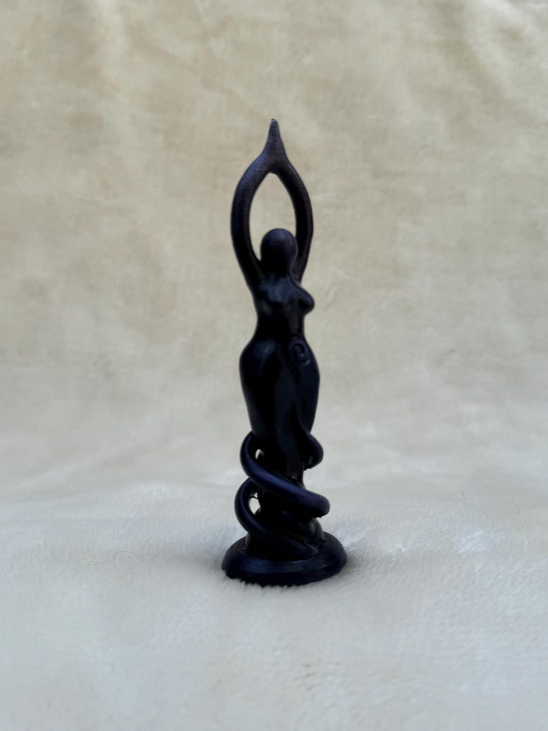 May include: Black figurine of a woman standing with her arms raised above her head, with a snake wrapped around her legs. The figurine is on a black base.