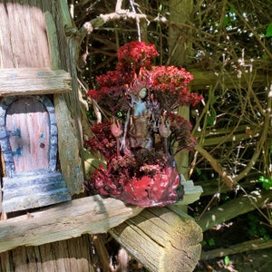 May include: A ceramic sculpture of a woman standing in a forest setting. The woman is surrounded by red and green foliage and flowers. The sculpture is painted in shades of brown, red, and green.