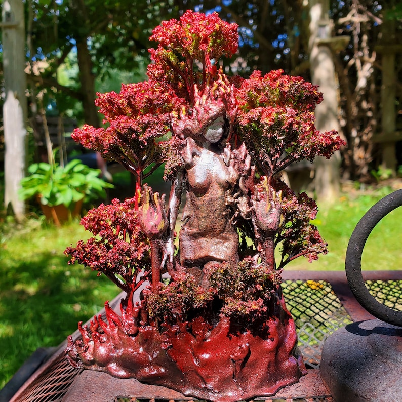 May include: A ceramic sculpture of a woman standing in a forest. The woman is painted in shades of silver and brown, and the trees are painted in shades of red and green.