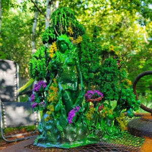 May include: A green resin sculpture of a woman standing in a forest. The sculpture is detailed with small trees, flowers, and leaves. The woman is wearing a flowing dress and has long hair.
