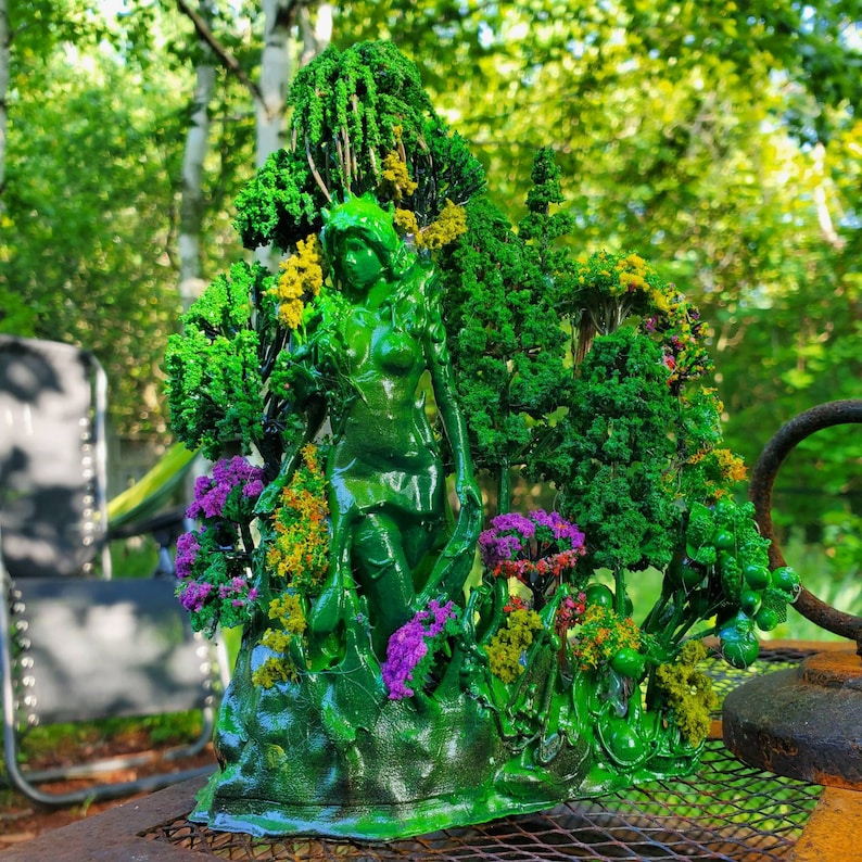May include: A green resin sculpture of a woman standing in a forest. The sculpture is detailed with small trees, flowers, and leaves. The woman is wearing a flowing dress and has long hair.