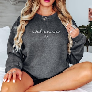 May include: A dark gray sweatshirt with the text "arbonne" and a small logo in white. The sweatshirt is worn by a person with long blonde hair.