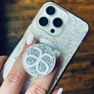 May include: A white phone case with a sparkly iridescent glitter design. A white phone grip with a silver glitter design is attached to the back of the phone case.