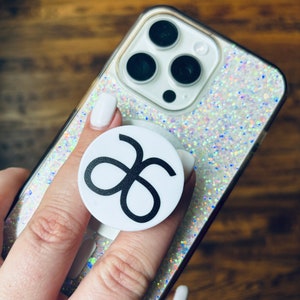 May include: A white phone case with a sparkly iridescent finish. A white pop socket with a black infinity symbol is attached to the back of the phone case.