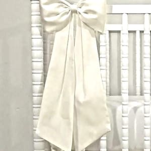 May include: A large, ivory-colored bow is tied to the head of a white crib. The bow's long, flowing tails cascade down the front of the crib. The crib has a white skirt and a white mattress.