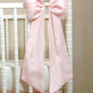 May include: A large, pearl blush taffeta bow adorns a white crib. The bow is tied in the center and has long, flowing tails. The crib has a matching pink skirt. The text "Pearl Blush Taffeta Bow" is at the top.