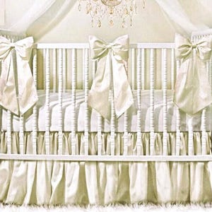 May include: A white crib with a cream-colored satin bow on each corner and a matching skirt. The crib has vertical slats and a soft, neutral color palette, creating a serene and elegant nursery setting.