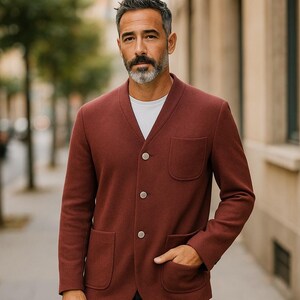 May include: A burgundy blazer with three buttons and two front pockets. The blazer is paired with a white t-shirt and dark blue jeans. The person is standing on a brick sidewalk.
