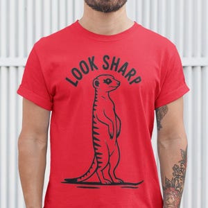 Puede incluir: Camiseta roja con la frase "LOOK SHARP" sobre una ilustración negra de un suricato. La persona que lleva la camiseta está de pie frente a una pared blanca con líneas verticales. La persona también lleva gafas y vaqueros.