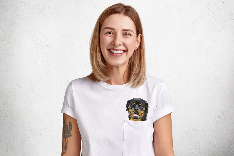 May include: White t-shirt with a pocket featuring a geometric illustration of a Rottweiler dog. The dog has black and brown fur with a yellow mouth. The person wearing the shirt is smiling.