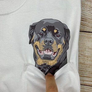May include: White t-shirt featuring a smiling Rottweiler dog graphic on the pocket. The dog has black, brown, and tan fur with a happy expression. The shirt is made of a soft, white fabric.
