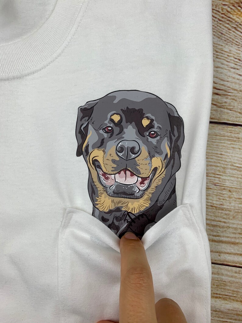 May include: White t-shirt featuring a smiling Rottweiler dog graphic on the pocket. The dog has black, brown, and tan fur with a happy expression. The shirt is made of a soft, white fabric.