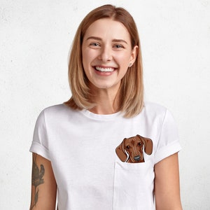 Puede incluir: Una mujer con una camiseta blanca con una ilustración de un perro salchicha marrón en el bolsillo. Ella está sonriendo y mirando a la cámara.