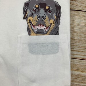 May include: White t-shirt with a pocket featuring a smiling Rottweiler dog graphic. The dog has black, tan, and brown fur, with a happy expression. The pocket is a light gray color.