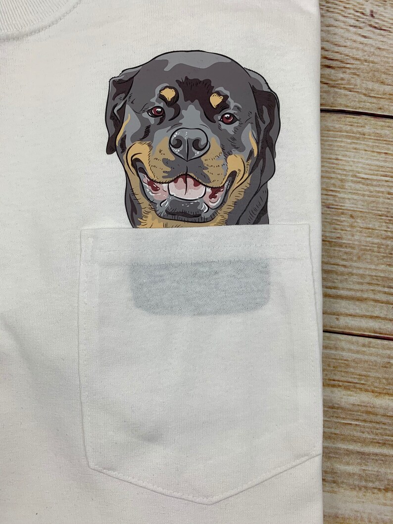May include: White t-shirt with a pocket featuring a smiling Rottweiler dog graphic. The dog has black, tan, and brown fur, with a happy expression. The pocket is a light gray color.