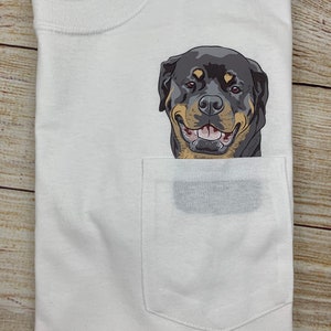 May include: White t-shirt with a pocket featuring a smiling Rottweiler dog graphic. The dog has black, tan, and brown fur. The shirt is folded on a wooden surface.