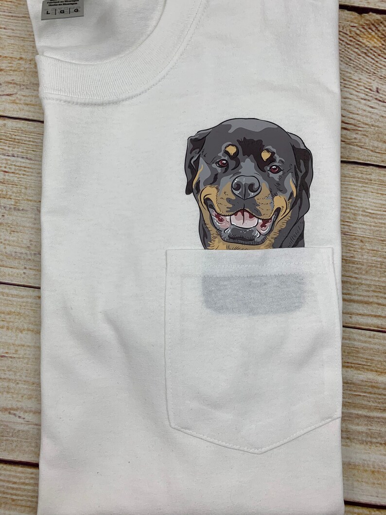 May include: White t-shirt with a pocket featuring a smiling Rottweiler dog graphic. The dog has black, tan, and brown fur. The shirt is folded on a wooden surface.