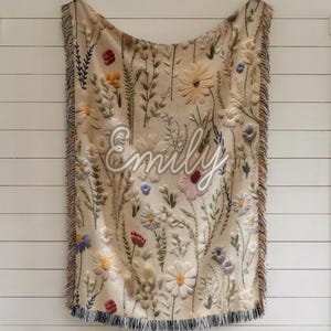 May include: Beige throw blanket with embroidered floral design and the name "Emily" in white script. The blanket features a variety of colorful flowers and foliage, with a fringed edge. The blanket is hanging on a white wall.