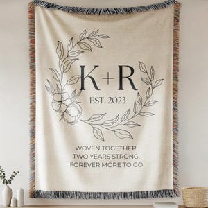 May include: Cream-colored woven throw blanket with black fringe and a floral wreath design. The blanket features the initials "K+R" and the year "2023", along with the text "WOVEN TOGETHER, TWO YEARS STRONG, FOREVER MORE TO GO".