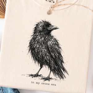 May include: An ivory t-shirt featuring a detailed black ink illustration of a crow standing on the ground. Below the bird, the text reads in my crone era. The shirt has a Comfort Colors brand label at the neckline and a round crew neck collar.