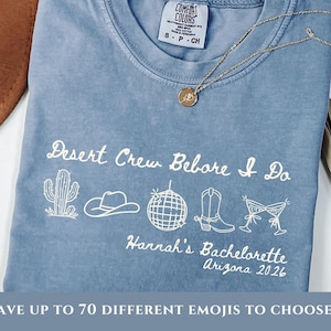 May include: A blue jean Comfort Colors t-shirt with the text "Desert Crew Before I Do" and "Hannah's Bachelorette Arizona 2026" in white. The shirt features cactus, cowboy hat, disco ball, boots, and cocktail glass graphics. A gold necklace is draped across the shirt.
