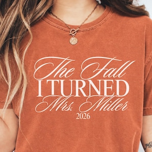 May include: A rust-colored short-sleeve t-shirt featuring white text that reads The Fall I TURNED Mrs. Miller 2026. The shirt has a crew neckline and a slightly faded, vintage-inspired appearance. The text is printed in a mix of script and serif fonts.
