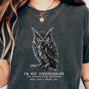 Puede incluir: Una camiseta de manga corta en color gris carb&oacute;n con una ilustraci&oacute;n detallada en l&iacute;neas blancas de un b&uacute;ho posado en una rama. Debajo del b&uacute;ho, el texto dice: I'M NOT OVERTHINKING I'm pre-solving problems that don't exist yet. Tiene cuello redondo.