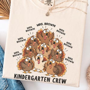 Puede incluir: Una camiseta de color marfil con un gráfico de varios pavos con los ojos muy abiertos y el pico abierto. Cada pavo está rodeado por el texto "MRS. BROWN!" y la parte inferior de la camiseta muestra el texto "KINDERGARTEN CREW" en letras negras.