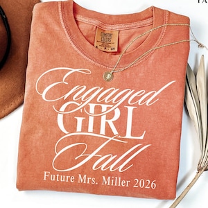 May include: A terracotta-colored short-sleeve t-shirt featuring white screen-printed text that reads Engaged GIRL Fall and Future Mrs. Miller 2026. The shirt has a crew neckline and a Comfort Colors brand label visible on the inside collar area.