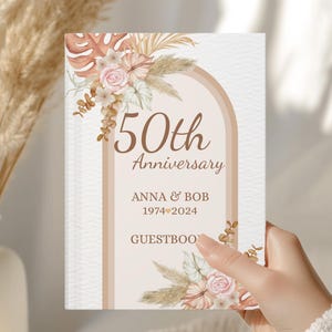 Puede incluir: Un libro de visitas blanco con el texto "50th Anniversary" y los nombres "ANNA & BOB 1974-2024". La portada presenta un diseño floral con acentos beige, rosa y marrón. El libro se sostiene en una mano.