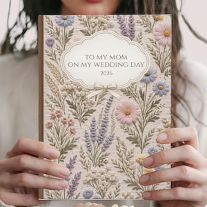 May include: A floral-patterned journal with the text "TO MY MOM ON MY WEDDING DAY 2026" in a decorative frame. The cover features a beige background with embroidered-style flowers in pink, purple, and blue. The word "FRONT" is at the bottom.