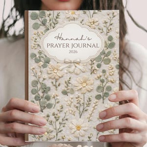 May include: A floral prayer journal with the text "Hannah's Prayer Journal 2026" in an ornate frame. The cover features a cream-colored background with embroidered flowers and greenery. The word "FRONT" is at the bottom.