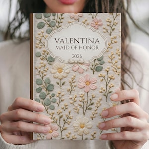 May include: A journal with a floral design and the text "VALENTINA MAID OF HONOR 2026". The cover features embroidered flowers in pink, cream, and green, with a cream-colored background. The journal has a brown spine.