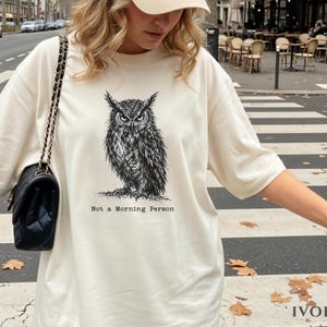 Puede incluir: Una camiseta color marfil con una ilustraci&oacute;n detallada en tinta negra de un b&uacute;ho con mirada intensa. Debajo del b&uacute;ho, el texto "Not a Morning Person" aparece en una fuente estilo m&aacute;quina de escribir. Tiene mangas cortas y cuello redondo.