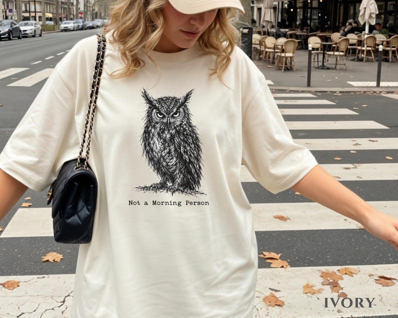 Puede incluir: Una camiseta color marfil con una ilustraci&oacute;n detallada en tinta negra de un b&uacute;ho con mirada intensa. Debajo del b&uacute;ho, el texto "Not a Morning Person" aparece en una fuente estilo m&aacute;quina de escribir. Tiene mangas cortas y cuello redondo.