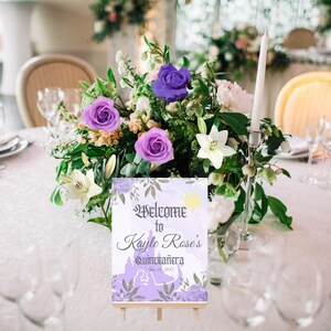 Puede incluir: Un cartel de bienvenida morado y blanco con una silueta de castillo y el texto "Bienvenido a la Quincea&ntilde;era de Kayle Rose" en un caballete de madera. El cartel est&aacute; rodeado de un arreglo floral con flores moradas, blancas y verdes.