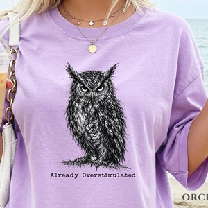 Puede incluir: Una camiseta de manga corta de color morado claro con una ilustraci&oacute;n detallada en blanco y negro estilo tinta de un b&uacute;ho. Debajo del b&uacute;ho, el texto "Already Overstimulated" aparece en una fuente de estilo m&aacute;quina de escribir.