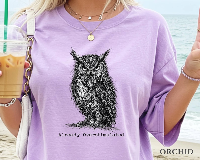 Puede incluir: Una camiseta de manga corta de color morado claro con una ilustraci&oacute;n detallada en blanco y negro estilo tinta de un b&uacute;ho. Debajo del b&uacute;ho, el texto "Already Overstimulated" aparece en una fuente de estilo m&aacute;quina de escribir.