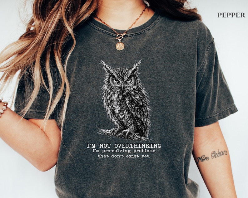 Puede incluir: Una camiseta de manga corta en color gris carb&oacute;n con una ilustraci&oacute;n detallada en l&iacute;neas blancas de un b&uacute;ho posado en una rama. Debajo del b&uacute;ho, el texto dice: I'M NOT OVERTHINKING I'm pre-solving problems that don't exist yet. Tiene cuello redondo.