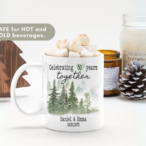Może przedstawiać: Biały ceramiczny kubek z uchwytem, wypełniony piankami marshmallow. Kubek przedstawia scenę leśną i napis "Celebrating 50 years together" z imionami pary i datą rocznicy. Kubek jest bezpieczny dla gorących i zimnych napoj&oacute;w.