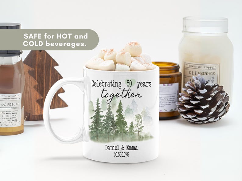 Może przedstawiać: Biały ceramiczny kubek z uchwytem, wypełniony piankami marshmallow. Kubek przedstawia scenę leśną i napis "Celebrating 50 years together" z imionami pary i datą rocznicy. Kubek jest bezpieczny dla gorących i zimnych napoj&oacute;w.