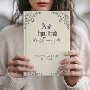 May include: A cream-colored book with the text "And they lived Happily ever after" in elegant script. The book is personalized with the names "ABIGAIL & FRANK" and the date "03-17-24". Decorative floral corner accents frame the text.