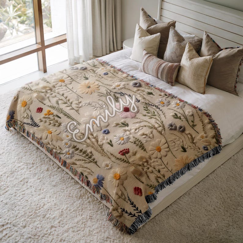 May include: Beige throw blanket with embroidered floral design, including daisies and other wildflowers. The blanket has a fringed edge and the name "Emily" in cursive. The blanket is on a bed with pillows.